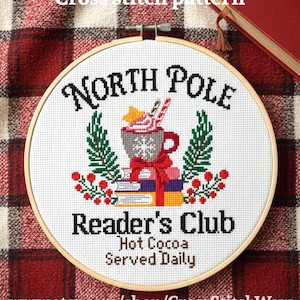 May include: A cross-stitch pattern in a wooden hoop features the words "North Pole Reader's Club" with a hot cocoa design. The design includes fir trees, berries, and the text "Hot Cocoa Served Daily". The background is a red and white plaid.
