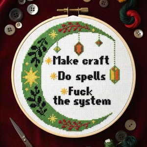 May include: Cross-stitch embroidery in a wooden hoop with the text "Make craft, Do spells, Fuck the system." The design features a green crescent moon with stars and decorative elements. Buttons, scissors, and thread are scattered around.