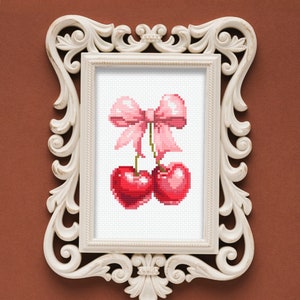 May include: A white ornate frame with a cross-stitch design of two red cherries with a pink bow on top. The cherries are on a white background.