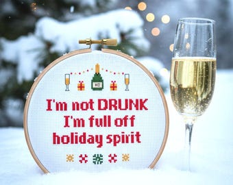 May include: A cross-stitch design in a wooden hoop with the text "I'm not DRUNK I'm full off holiday spirit." The design includes champagne glasses, gifts, and a champagne bottle. A champagne flute is next to the hoop, set in snow.