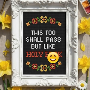 May include: A framed cross-stitch with the text "THIS TOO SHALL PASS BUT LIKE HOLY F**K" and a smiling emoji. The white frame has ornate detailing. Yellow daffodils and daisies surround the frame.