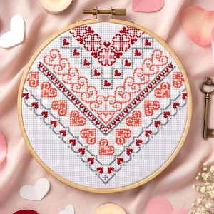 Könnte beinhalten: Ein Kreuzstich-Herz-Design in einem hölzernen Stickrahmen. Das Design zeigt rote, korallenfarbene und graue Muster, darunter Herzen und geometrische Formen. Der Hintergrund umfasst Kerzen, Rosenblätter und herzförmige Ausschnitte.