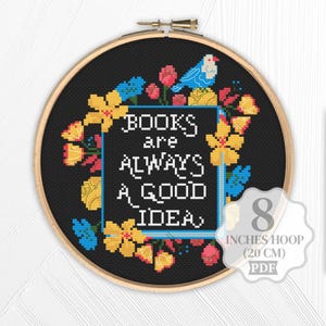 Op de afbeelding: Kruissteekborduurwerk in een houten hoepel. Het ontwerp toont de tekst "BOOKS are ALWAYS A GOOD IDEA" in het wit, omringd door gele, roze en blauwe bloemelementen en een blauwe vogel. De hoepel heeft een diameter van 20 cm.