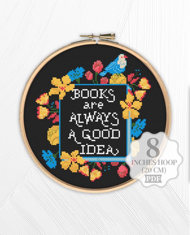 Op de afbeelding: Kruissteekborduurwerk in een houten hoepel. Het ontwerp toont de tekst "BOOKS are ALWAYS A GOOD IDEA" in het wit, omringd door gele, roze en blauwe bloemelementen en een blauwe vogel. De hoepel heeft een diameter van 20 cm.