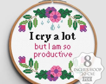May include: Cross stitch pattern featuring a floral wreath with pink and purple flowers and green leaves. The text "I cry a lot but I am so productive" is stitched in black thread on a white background. The pattern is designed for an 8-inch hoop.