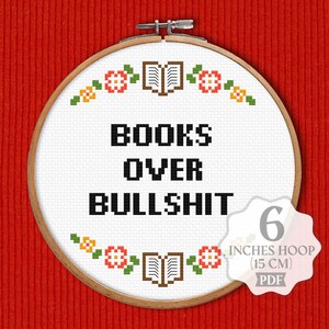 K&ouml;nnte beinhalten: Ein wei&szlig;er Stickrahmen mit einem Kreuzstich-Design, das "BOOKS OVER BULLSHIT" lautet. Das Design ist von einem Rand aus kleinen Blumen und B&uuml;chern umgeben. Der Rahmen hat einen Durchmesser von 15 cm.