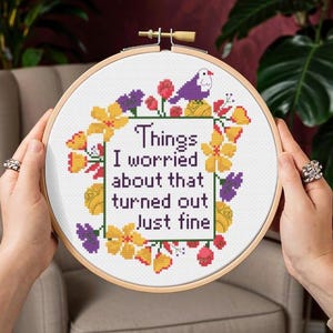 Op de afbeelding: Een kruissteek borduurring met de tekst "Things I worried about that turned out Just fine." Het ontwerp bevat een bloemenrand met gele, paarse en rode bloemen en een kleine paarse vogel. De ring wordt door twee handen vastgehouden.