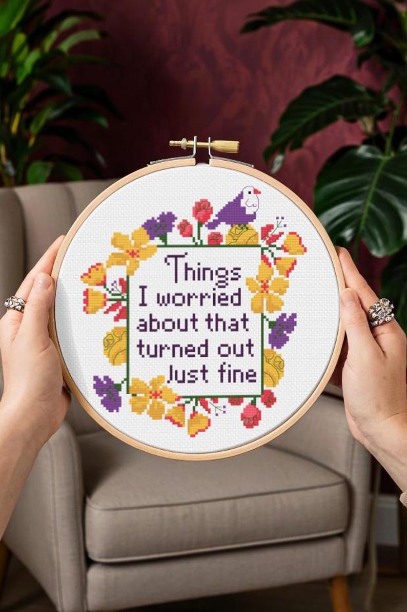 Op de afbeelding: Een kruissteek borduurring met de tekst "Things I worried about that turned out Just fine." Het ontwerp bevat een bloemenrand met gele, paarse en rode bloemen en een kleine paarse vogel. De ring wordt door twee handen vastgehouden.