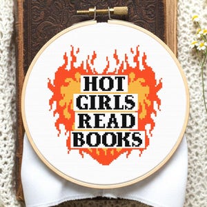 Op de afbeelding: Een borduurring met de tekst "HOT GIRLS READ BOOKS" in zwart en oranje, omgeven door een vlammenontwerp. De ring staat op een open, antiek boek en een witte, getextureerde stof.