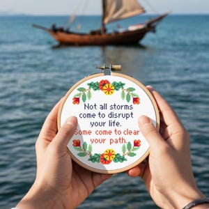 Op de afbeelding: Een ronde, geborduurde hoepel met de tekst "Not all storms come to disrupt your life. Some come to clear your path." Het ontwerp bevat bloemaccenten in rood, geel en groen. Een zeilboot is op de achtergrond te zien.
