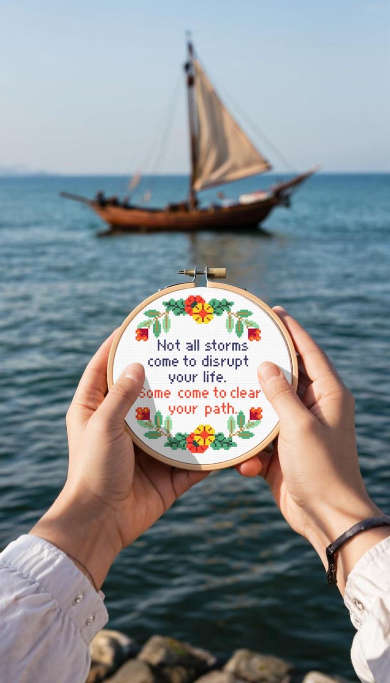 Op de afbeelding: Een ronde, geborduurde hoepel met de tekst "Not all storms come to disrupt your life. Some come to clear your path." Het ontwerp bevat bloemaccenten in rood, geel en groen. Een zeilboot is op de achtergrond te zien.
