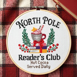 Op de afbeelding: Een ronde houten borduurring met witte stof en een kruissteekontwerp. De tekst luidt North Pole Reader's Club Hot Cocoa Served Daily. De afbeelding toont een mok cacao, boeken, dennentakken en rode bessen.