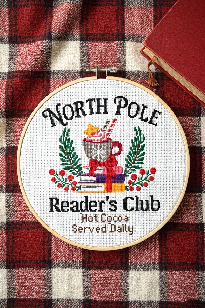 Op de afbeelding: Een ronde houten borduurring met witte stof en een kruissteekontwerp. De tekst luidt North Pole Reader's Club Hot Cocoa Served Daily. De afbeelding toont een mok cacao, boeken, dennentakken en rode bessen.