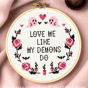 May include: A cross-stitch pattern in a wooden hoop with the text "Love Me Like My Demons Do." The design includes pink ghosts, hearts, roses, and candles. The Etsy shop name is visible at the bottom.