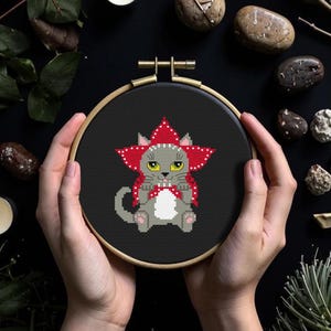 May include: A cross-stitch embroidery hoop featuring a gray cat wearing a red flower-shaped hat. The cat has yellow eyes and is seated on a black background. The embroidery hoop is held in hands, with a black background and decorative elements.