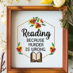 May include: A framed cross-stitch design with a white background and a hexagonal border of yellow and orange flowers. The text reads "Reading because murder is wrong" with a book illustration below.