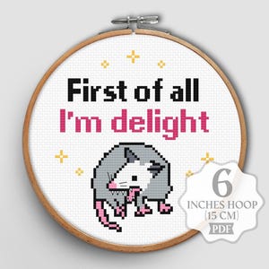 Puede incluir: Un patrón de punto de cruz en un bastidor de madera de 15 cm con una zarigüeya pixelada de nariz y patas rosas. El texto First of all I'm delight aparece en una fuente pixelada negra y rosa, rodeado de pequeñas estrellas amarillas bordadas.