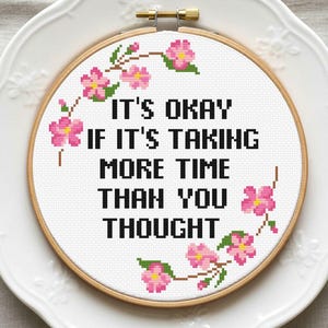 May include: Cross-stitch embroidery in a wooden hoop with the text "IT'S OKAY IF IT'S TAKING MORE TIME THAN YOU THOUGHT." Pink flowers and green leaves decorate the design. The hoop is on a white plate.