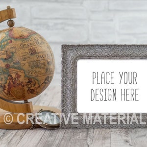 Könnte beinhalten: Ein silberner, verzierter Bilderrahmen mit weißem Hintergrund und dem Text "PLACE YOUR DESIGN HERE". Der Rahmen steht auf einer Holzoberfläche neben einem antiken Globus und einem Kompass.