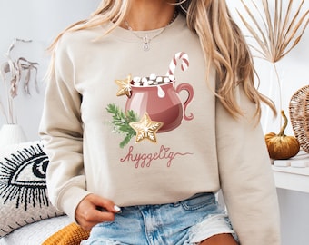 May include: Beige sweatshirt featuring a festive graphic of a mug of hot cocoa with marshmallows, a candy cane, and gingerbread stars. The word "hyggelig" is written in script below the mug. A cozy, seasonal design.