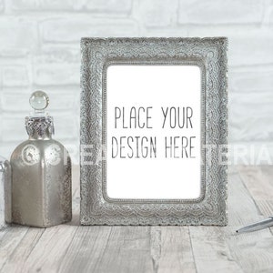 Könnte beinhalten: Ein silberner Bilderrahmen mit einem verzierten Design steht auf einer Holzoberfläche mit zwei silbernen Flaschen und einem silbernen Stift. Der Rahmen hat einen weißen Hintergrund mit dem Text "Place your design here".