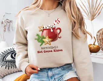 May include: Beige sweatshirt with a festive graphic. The design features a red mug of hot cocoa with marshmallows, a candy cane, and gingerbread stars. The text "Warm Wishes & Hot Cocoa Kisses" is printed below the graphic. A cozy winter-themed apparel item.