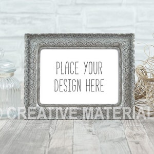 Könnte beinhalten: Ein silberner, verzierter Bilderrahmen mit einem weißen Passepartout und dem Text "Place your design here" auf einem weiß getünchten Holzgrund.