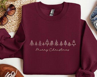 May include: A maroon sweatshirt with a white line drawing of Christmas trees and the words "Merry Christmas" in a script font. The sweatshirt is folded on a woven mat, with sunglasses and a shoe visible.