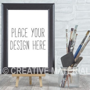 May include: A black picture frame with a white background and the text "PLACE YOUR DESIGN HERE" is on a small wooden easel. The frame is in front of a white brick wall. A glass jar with paintbrushes is on a wooden surface.