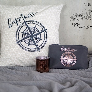Könnte beinhalten: Zwei dekorative Kissen mit einem Kompass-Design und dem Wort "happiness" darauf gedruckt. Die Kissen liegen auf einer grauen Decke mit einem Glas-Kerzenhalter.