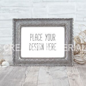 Puede incluir: Un marco de fotos plateado adornado con un fondo blanco y el texto "PLACE YOUR DESIGN HERE". El marco está sobre una superficie de madera clara con una planta de hierba seca y conchas.