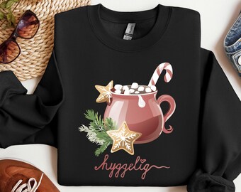 May include: Black sweatshirt featuring a festive design. A red mug overflowing with marshmallows and a candy cane is surrounded by gingerbread cookies and greenery. The word "hyggelig" is written in cursive.