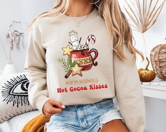 May include: Beige sweatshirt featuring a festive graphic of a mug of hot cocoa with marshmallows, a candy cane, and star-shaped cookies. The text "warm wishes & Hot Cocoa Kisses" is printed below the graphic. The sweatshirt is a cozy, casual style.