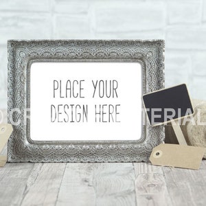 Puede incluir: Un marco de fotos plateado adornado con un paspartú blanco y el texto "PLACE YOUR DESIGN HERE" en su interior. El marco está sobre una superficie de madera con un pequeño letrero de pizarra, una etiqueta marrón y un rollo de cuerda.