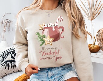 May include: Beige sweatshirt with a festive graphic. The design features a maroon mug filled with marshmallows, a candy cane, and gingerbread stars. Text reads "It's the most Wonderful time of the Year".