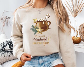May include: Beige sweatshirt with a festive graphic. The design features a brown mug of hot chocolate with marshmallows, a candy cane, and star-shaped cookies. Text reads "It's the most Wonderful time of the Year."