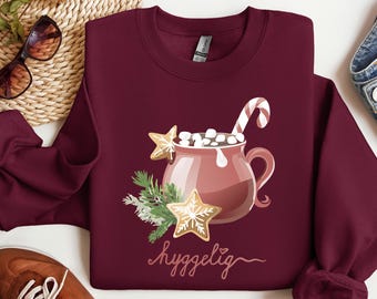 May include: Burgundy sweatshirt featuring a festive illustration of a mug of hot cocoa with marshmallows, a candy cane, and gingerbread cookies. The design includes evergreen sprigs and the word "hyggelig" in script. A cozy holiday-themed apparel item.