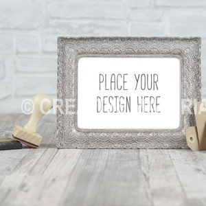 Könnte beinhalten: Ein silberner, verzierter Bilderrahmen mit weißem Hintergrund und dem Text "Place your design here". Der Rahmen steht auf einer hellbraunen Holzoberfläche mit einem Holzstempel und zwei braunen Papieretiketten.
