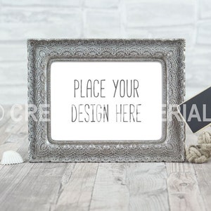 Puede incluir: Un marco de fotos plateado adornado con un paspartú blanco y el texto "PLACE YOUR DESIGN HERE" sobre un fondo de madera blanqueada. También hay un pequeño letrero de pizarra y un trozo de cuerda en la imagen.