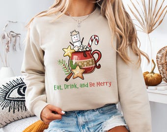 May include: Beige sweatshirt with a festive graphic. The design features a red mug filled with marshmallows, a candy cane, and gingerbread cookies. The text "Eat, Drink, and Be Merry" is printed below the graphic. A cozy holiday theme.