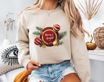May include: A beige sweatshirt with a Christmas-themed design. The graphic includes a red and gold circular ornament with the text "Merry & Bright!" surrounded by pine branches and baubles. A festive, cosy top.