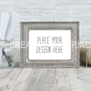 Könnte beinhalten: Ein silberner, verzierter Bilderrahmen mit einem weißen Passepartout und dem Text "Place your design here" auf einem weiß getünchten Holzgrund.