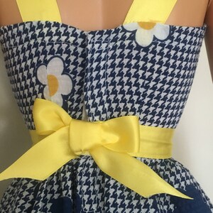 May include: A blue and white gingham dress with a yellow bow and white flowers. The dress has yellow straps.
