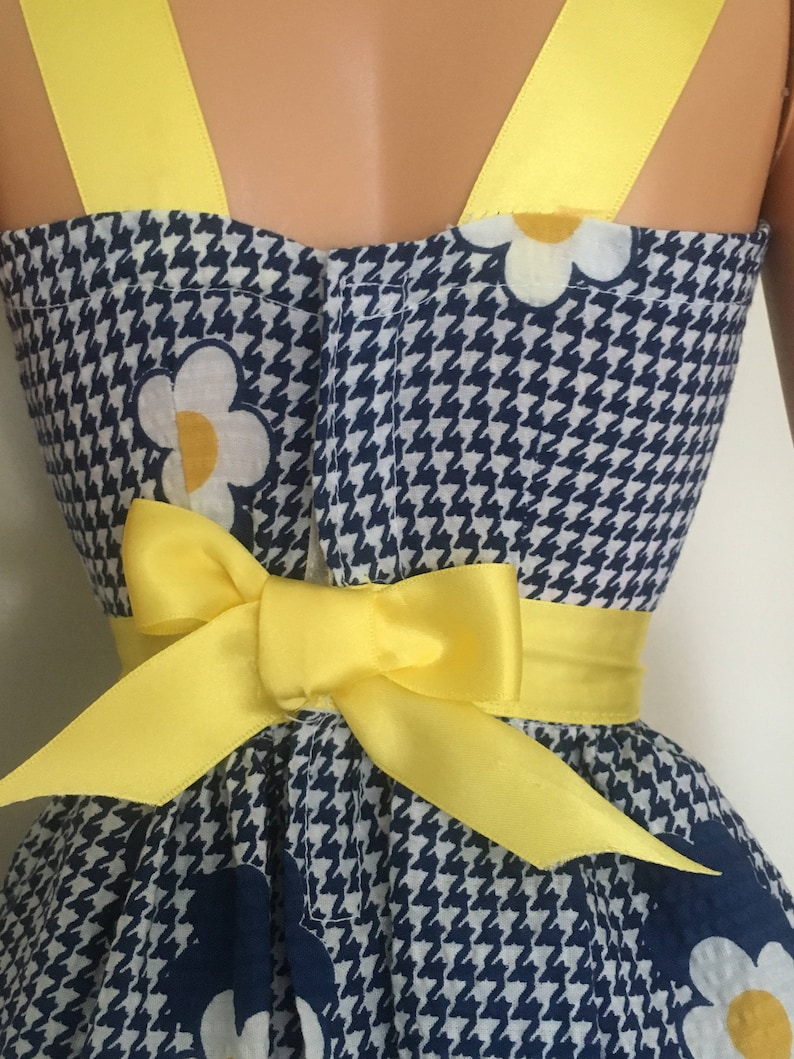May include: A blue and white gingham dress with a yellow bow and white flowers. The dress has yellow straps.