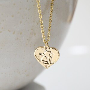 May include: A gold necklace with a textured heart pendant. The necklace is hanging from the edge of a white ceramic bowl.