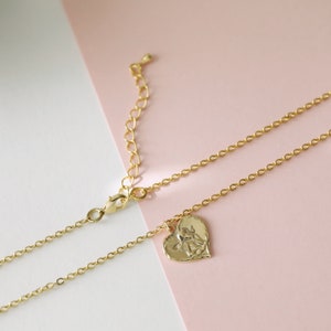 May include: A gold chain necklace with a heart-shaped pendant featuring an angel design. The necklace is on a pink and white background.