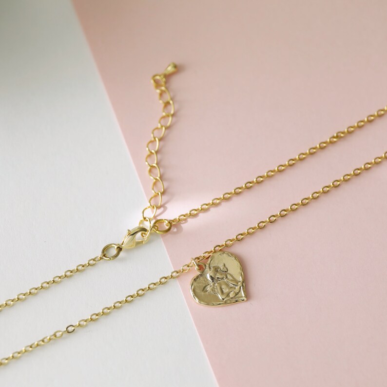 May include: A gold chain necklace with a heart-shaped pendant featuring an angel design. The necklace is on a pink and white background.