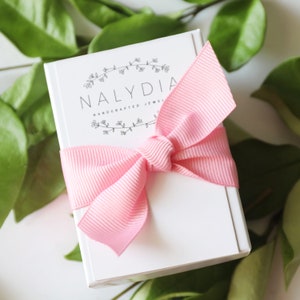 May include: A white gift box with a pink ribbon tied in a bow. The box has the text "NALYDIA HANDCRAFTED JEWELRY" printed on the front.