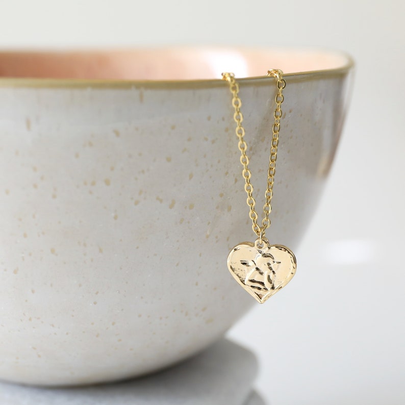 May include: A gold necklace with a textured heart pendant. The necklace is hanging from the edge of a white ceramic bowl.