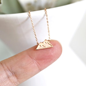 May include: A delicate gold-colored necklace featuring a mountain range pendant. The pendant is a small, triangular shape with etched mountain details. The chain is fine and the necklace is displayed against a white background.